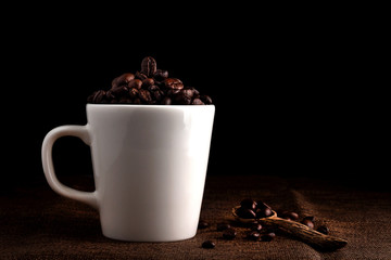 White cup full of coffee beans on the cloth sack in morning light and black background with copy space