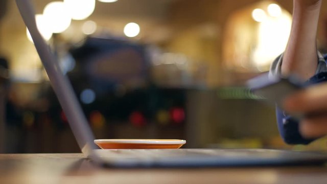 Profile close-up of laptop and hand lifting a cup of coffee, in coffee shop, blurred background. Somebody working, studying, writing. High quality 4K ProRes 422 HQ