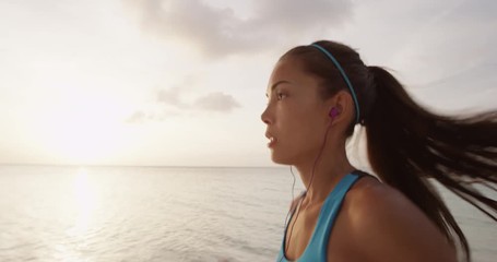 Determined female runner jogging at beach during sunset. Sporty young woman is listening music through earphones against sky wearing blue sports bra. SLOW MOTION, STEADICAM, RED EPIC. - Powered by Adobe