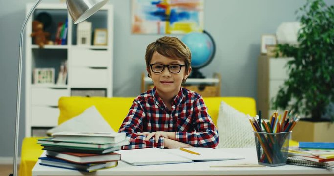 Portrait shot of the Caucasian cute small boy in glasses sitting at the desk with school stuff and textbooks in his room and smiling to the camera.