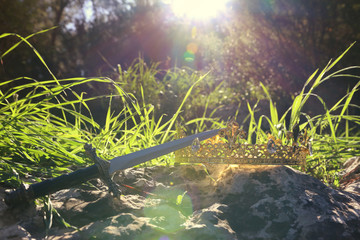 mysterious and magical photo of silver king crown and sword over the stone in the England woods or field landscape with light flare. Medieval period concept.