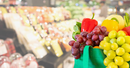 Fresh fruits and vegetables in reusable green shopping bag with supermarket grocery store blurred...