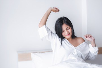 Beautiful young pretty Asian woman wake up and make happy smile with white shirt at the white bed in the morning.  