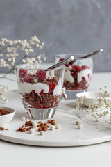 Two glasses of layered granola and yogurt with raspberries on a marble tray on a dark background