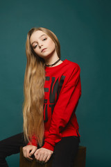 Stylish teen, beautiful young model girl with long blonde hair in black jeans and in fashionable red sweatshirt posing at the studio