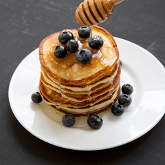 Pancakes with blueberries and honey on a white round plate over dark black background, side view. Close-up.