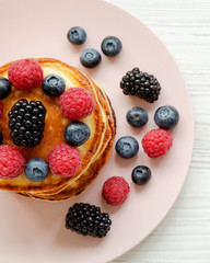 Pancakes with berries on a pink plate over white wooden surface, overhead view. From above, top view.