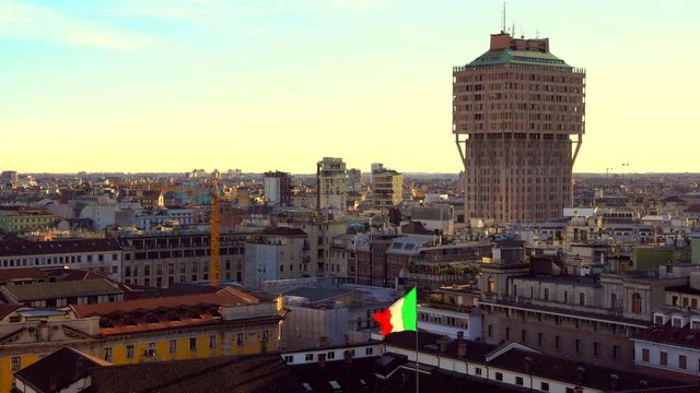 Milan skyline from the Duomo (Cathedral) roof, Italy. Panoramic view of Milan with sunset warm light. An Italian flag waving in the foreground.