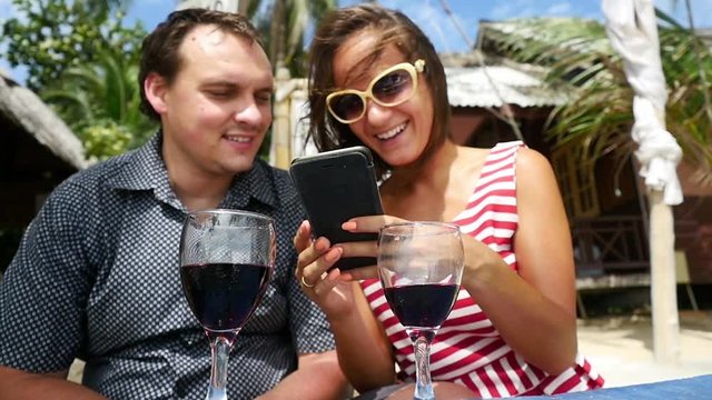 Young joyful couple drinks wine use smartphone having rest in outdoor beach cafe . slow motion. 1920x1080