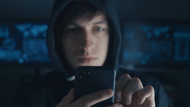 Dangerous Hacker in the hood uses a smartphone in a secret data center filled with screens