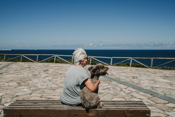 White hair middle-aged woman sitting in the company of her friendly dog ​​on a wooden bench overlooking the sea on a sunny and breezy summer morning. Lifestyle.