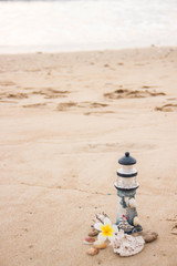 A small decorative lighthouse with a magnolia flower and shells is laid out on the sand overlooking the sea with a place for an inscription.