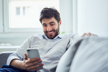 Cheerful young man using phone while relaxing on sofa after work