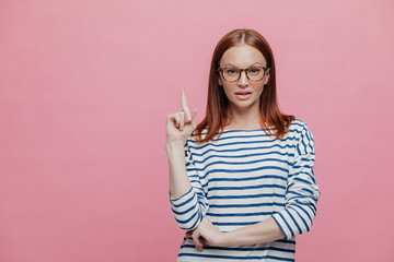 Photo of serious female shop assistant points fore finger upwards, shows blank space for your advertisement, demonstrates place for buying things with discounts, isolated over pink background