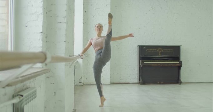 Graceful classic ballet dancer practicing battement soutenu exercise at barre during rehearsal in ballet studio. Ballerina performing different choreographic exercises and dance moves in dance studio.