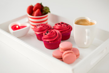 valentines day and sweets concept - close up of cupcakes with red buttercream frosting, macarons, heart shaped coffee cup, candle and strawberries on tray