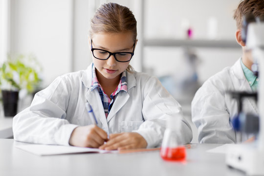 education, science and children concept - girl studying chemistry at school laboratory and writing to workbook
