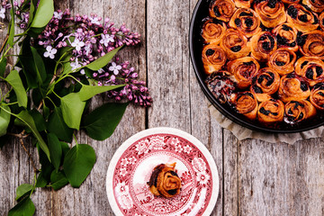 Homemade mini cherry rolls in a baking form and one roll on a plate with bouquet of lilac on vintage wooden table. Bun cake with cherry filling