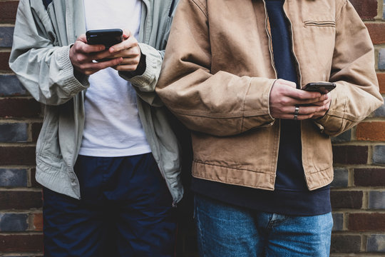 Mid section, two young men wearing casual jackets standing side by side against a brick wall, texting on their mobile phones.