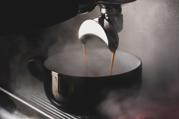 Close up of black mug on espresso machine, hot coffee pouring from spout.