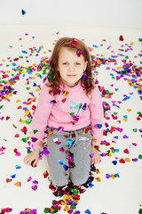 Obraz na płótnie Canvas Happy beautiful little girl enjoying colorful confetti surprise falling down, posing on white studio wall. Pretty girl celebrating her birthday party, having fun