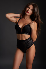 Stunning caucasian female model with dark hair and red lips in black underwear posing on dark grey background.