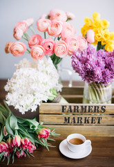 Beautiful spring flower set on the wooden table: fresh blossoming pink ranunculus, yellow Narcissus, purple and white hyacinths and magenta tulips in the wooden box and a cup of fresh espresso coffee
