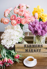 Beautiful spring flower set on the wooden table: fresh blossoming pink ranunculus, yellow Narcissus, purple and white hyacinths and magenta tulips in the wooden box and a cup of fresh espresso coffee