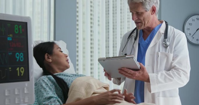 Older Male Doctor Going Over Chart With Female Patient Lying In Hospital Bed