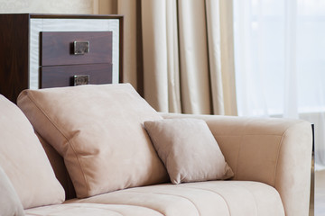 Close up of big and little pillows on beige sofa. Cozy place in living room with modern interior. Wooden nightstand with drawers on background.