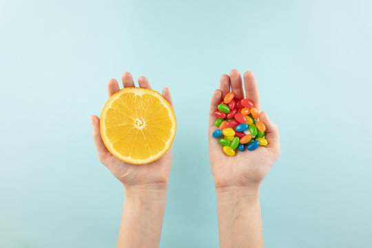 Vitamin pills and fresh orange in hands, top view. Female hands holding fruit in one hand and bunch of sweets in the other in simple background