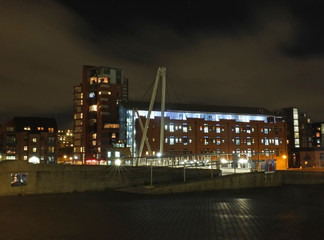 Obraz premium a cityscape view of the canal entrance to the clarence dock area of leeds with a pedestrian bridge crossing the water with reflections of lights and buildings against a night sky with clouds
