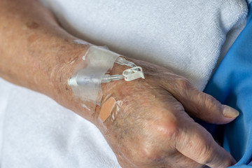 Hand of elderly patient with intravenous catheter for injection plug in hand during lying in hospital ward room (selective focus)
