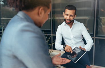 Fototapeta premium Two trendy businessmen pointing to tablet and talking to each other using hand gestures