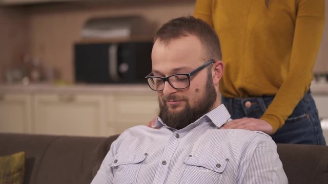 A bearded man sitting on the sofa and a young brunette standing behind him and massaging his shoulders. His eyes are closed and he throws back head a bit