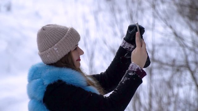 Young beautiful girl walking in the snowy winter forest. She's happy. A girl is taking pictures on a smartphone. Makes a selfie. Slow motion.