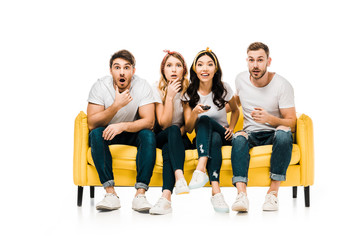 shocked young friends sitting on sofa with remote controller and looking at camera isolated on white