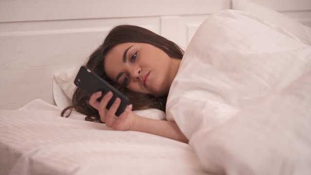 A closeup of a pretty long-haired brunette is lying sidelong in bed looking at the phone and scrolling it. She smiles a bit in the end