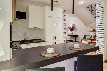 Stylish and modern interior of kitchen in beige, black and white colors. Two white cups on kitchen bar, two lamp over. Elements of stones, white wall and black chair around.