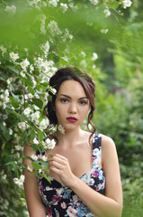 Young beautiful dark-haired woman in color dress with open shoulders in bright green blooming garden background