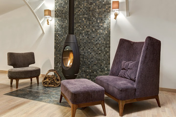 Stylish interior of living room with fireplace where fire is burning and basket with firewood near. Two armchair and little table. Room in gray, violet and white colors.