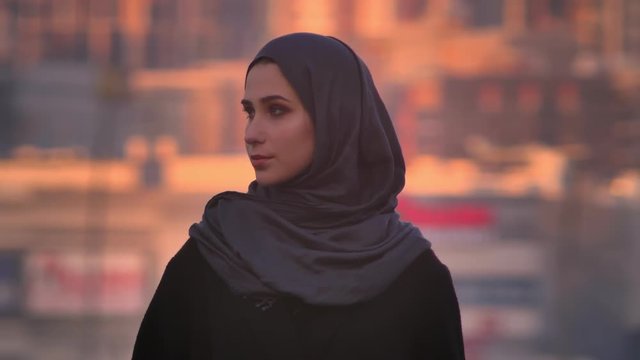 Closeup portrait of young attractive female in hijab looking confidently at camera with the evening city on the background
