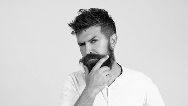 handsome bearded hipster man portrait with lush moustache and stylish hair on thoughtful face and white T-shirt touching long beard and thinking isolated. thoughtful young man standing by the wall.