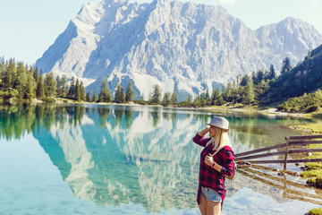 back view of blonde girl in hat with backpack standing near lake with sunlight