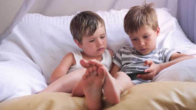 Two boys holding smartphone, tablet sitting on chair, focus on childrens feet. Alternative education concept. Preschollers with wireless gadget. Get bored background.