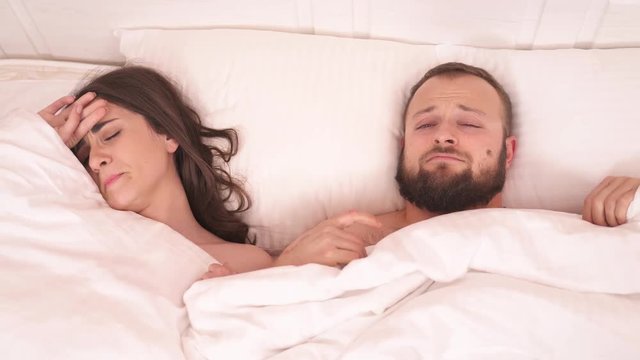A top view of white blanket on the bed taken away and a pretty brunette with a bearded man lying on the bed. They yawn, open and close eyes