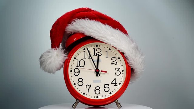 Christmas time concept. Santa Claus hat and big red clock ticking showing twelve hours, on grey background. Waiting for celebration. Time lapse