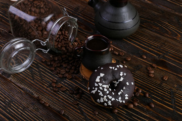 Donut with black icing and chocolate powder and an authentic cup of strong coffee. A can of coffee beans and poured grains.