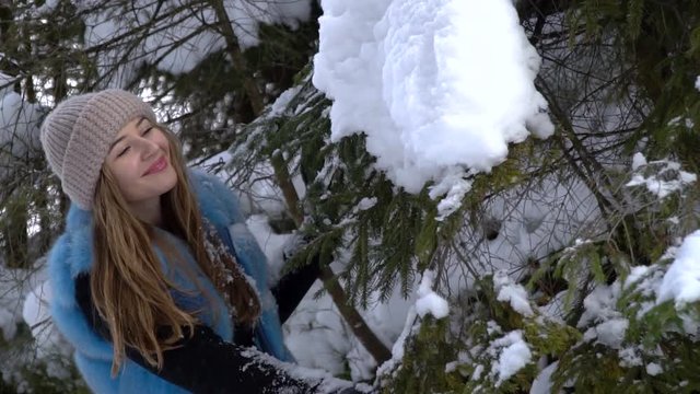 Young beautiful girl walking in the snowy winter forest. She is happy and calm. A girl stands near a large snow-covered spruce and carefully examines it. Slow motion. Full HD.