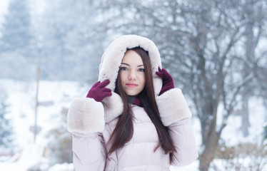 Portrait of young beautiful emotional woman in hooded down coat and gloves on snow covered garden background. Winter snowy landscape